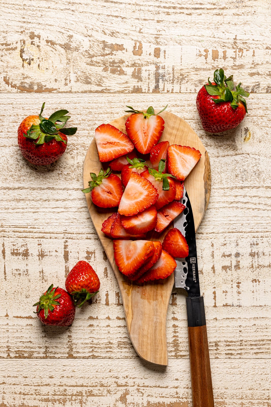 Strawberries sliced unto a wood cutting board on a white wood table with a small knife.