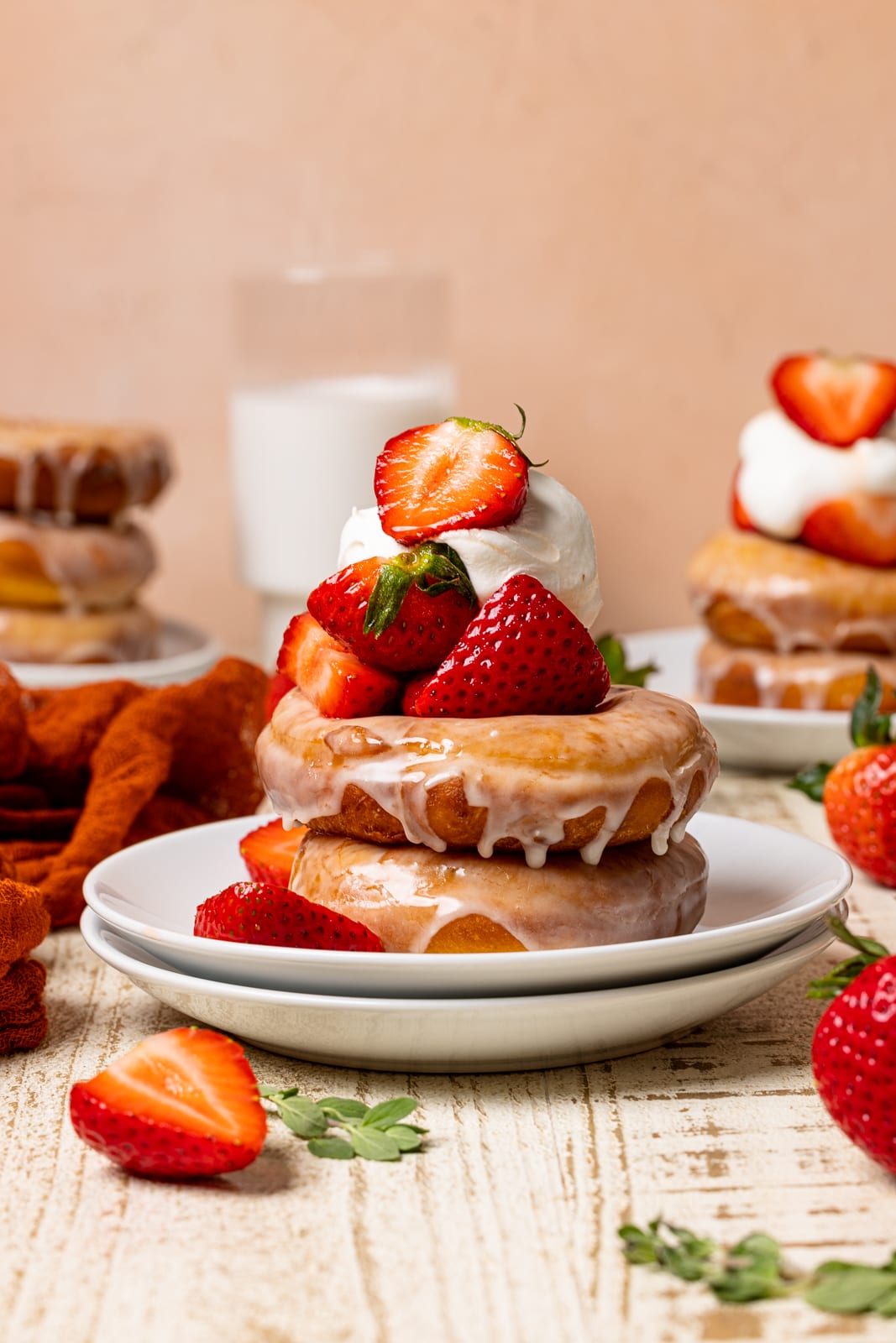Stacked donuts on two white plates with strawberries, whipped cream, and milk in the background on a white wood table.