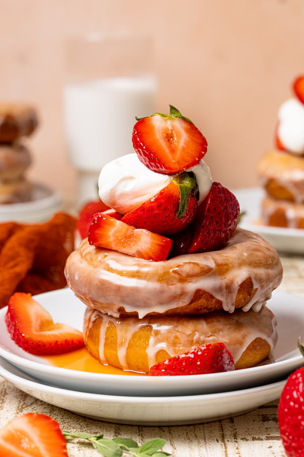 Stacked donuts on two white plates with strawberries, whipped cream, and milk in the background on a white wood table.
