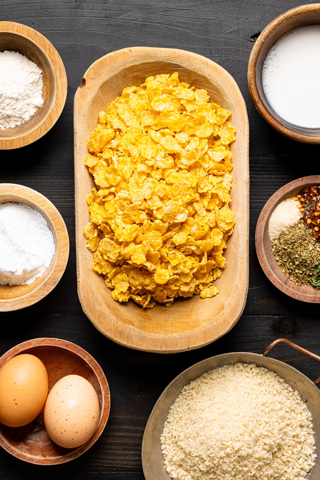 Ingredients on a black table including cornflakes, eggs, flour, breadcrumbs, herbs + seasonings, and milk.