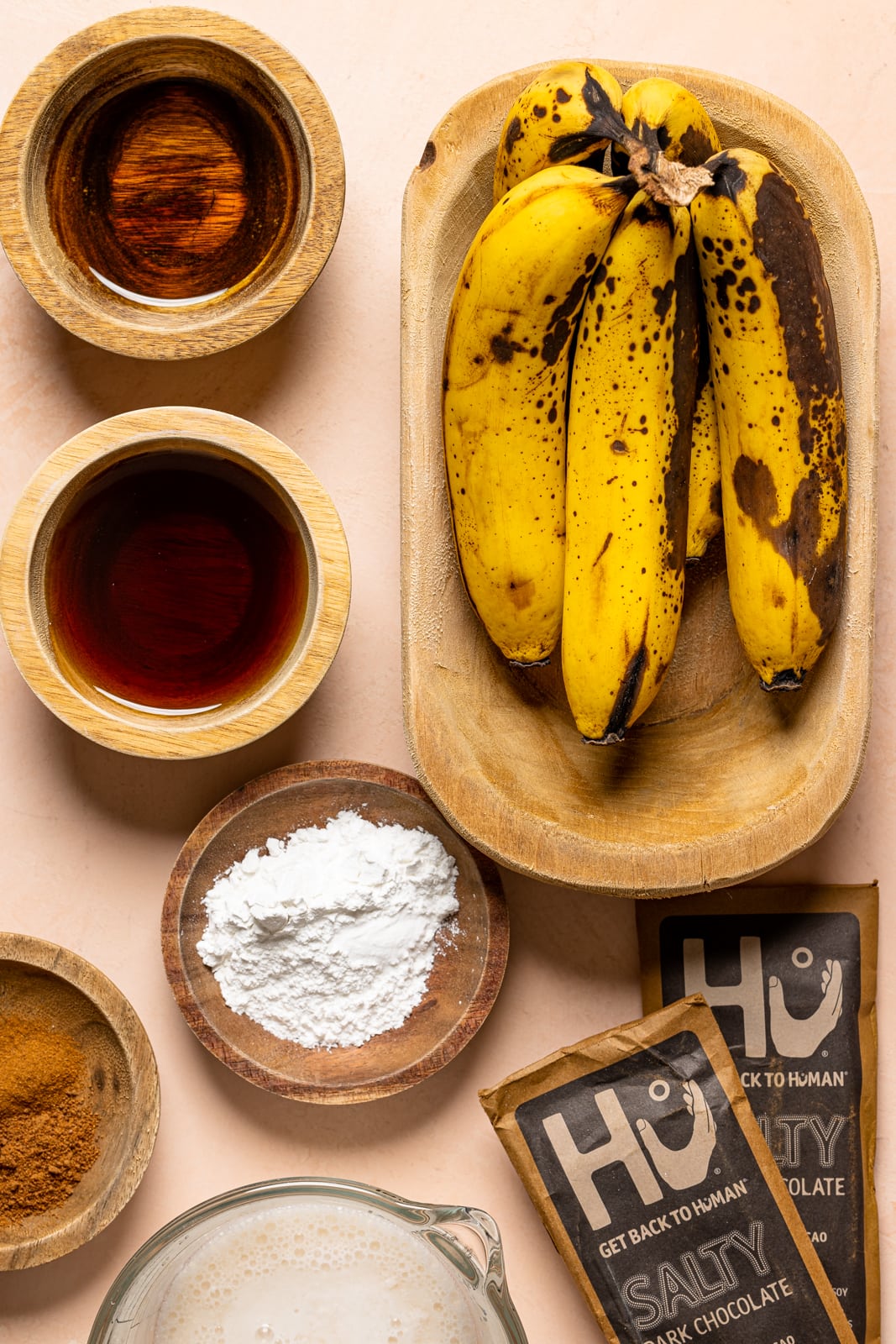 Ingredients on a peach table including bananas, maple syrup, chocolate, spices, milk, etc.