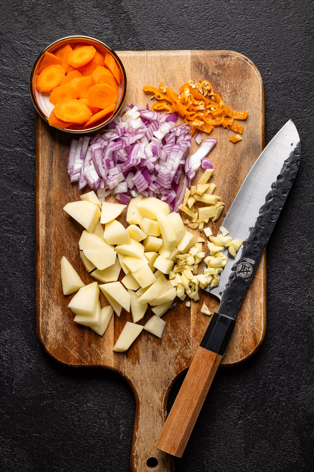 Chopped onions, potatoes, carrots, ginger, and peppers on a cutting board with a knife. 