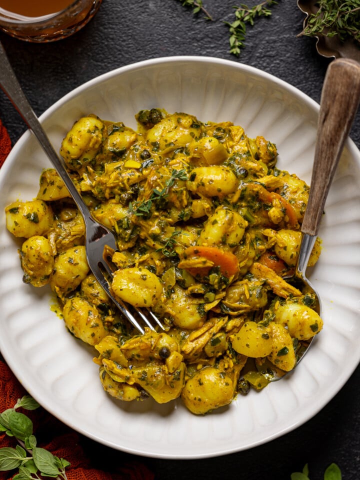 Bowl of curry chicken and gnocchi with a fork + spoon with a cup of juice.