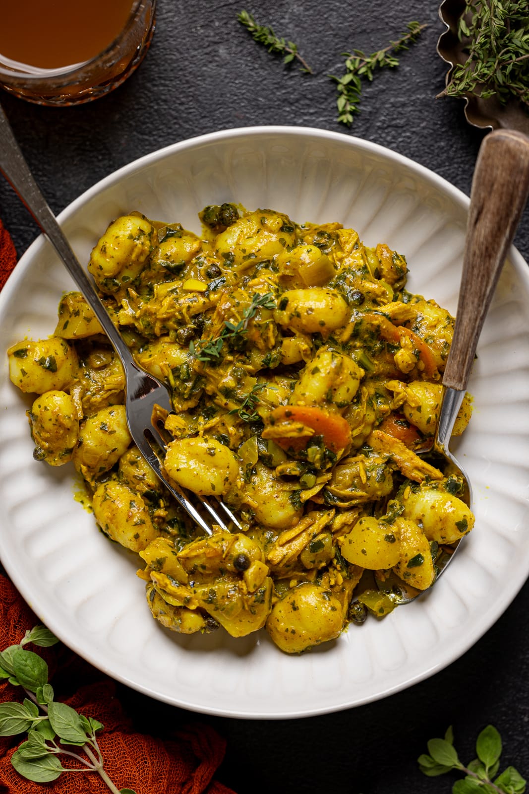 Bowl of curry chicken and gnocchi with a fork + spoon with a cup of juice.