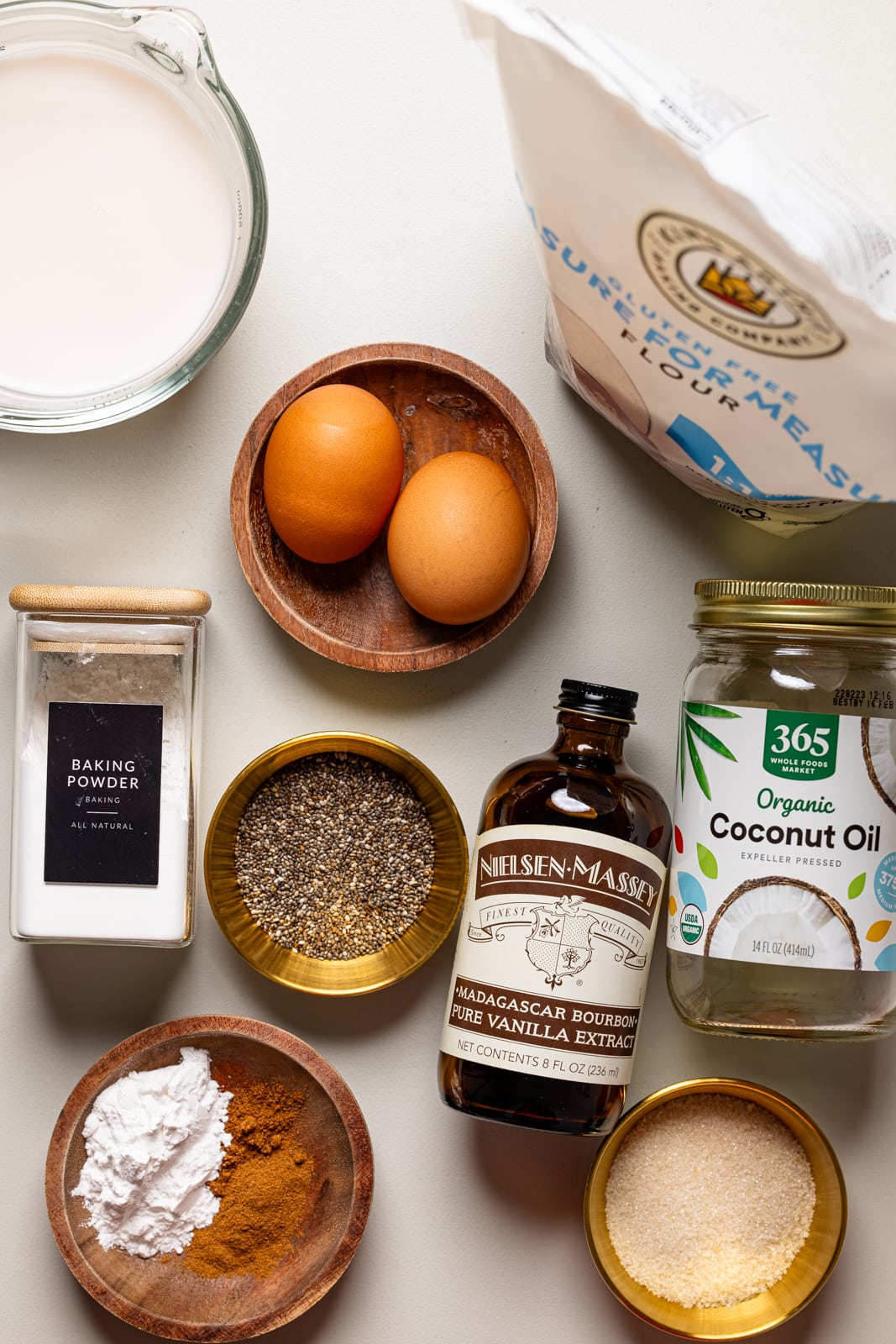 Ingredients on a grey table including gluten-free flour, eggs, vanilla, coconut oil, chia seeds, and spices.