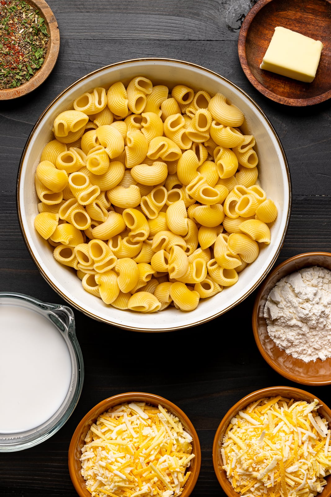 Ingredients on a black table including macaroni, milk, cheese, butter, flour, and herbs + seasonings.  