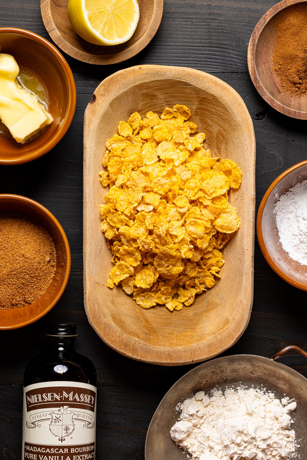 Ingredients on a black table including cornflakes, vanilla, butter, cinnamon, lemon, sugar, and flour. 