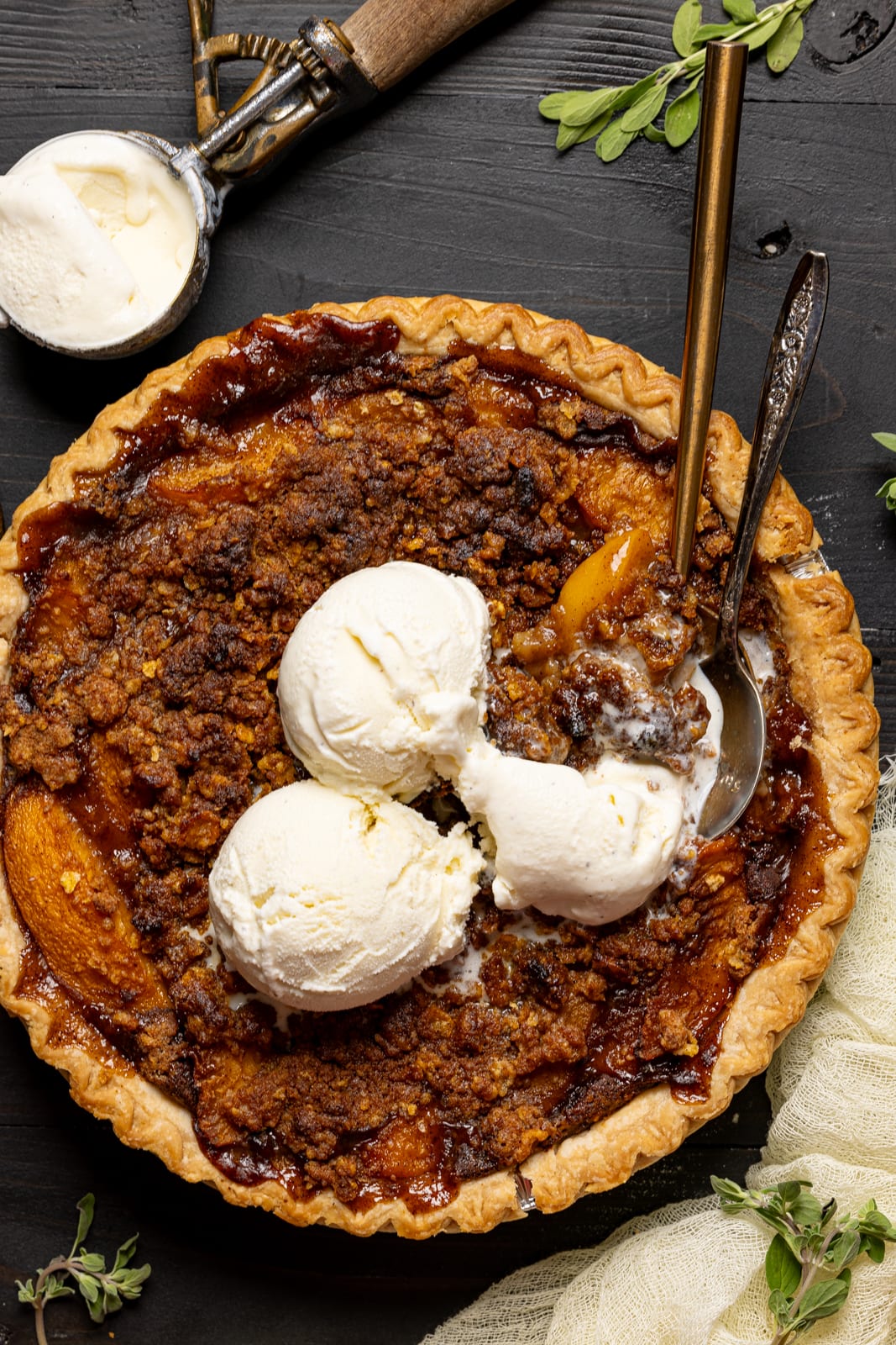 Pie on a black wood table with scoops of ice cream, spoons, and herbs. 