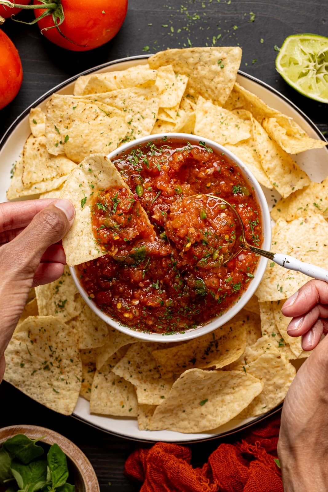 Salsa and chips with a spoon on a black table with tomatoes and lime.