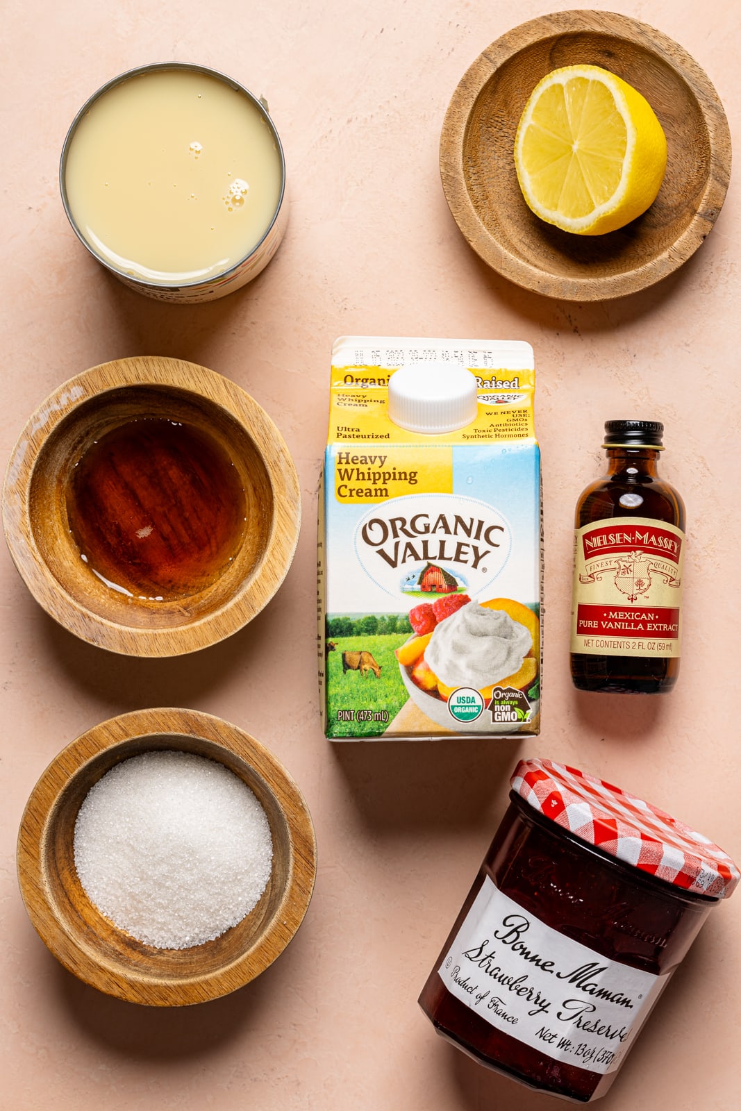 Ingredients on a peach table including heavy cream, jam, sugar, maple syrup, lemon, vanilla, and condensed milk.