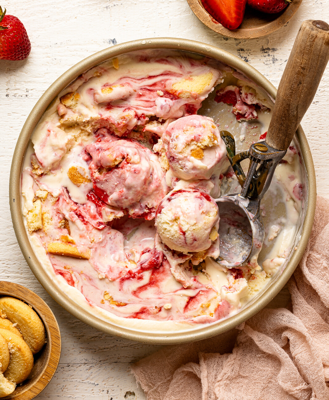 Ice cream in a container with scoops on a white wood table with strawberries and cookies.