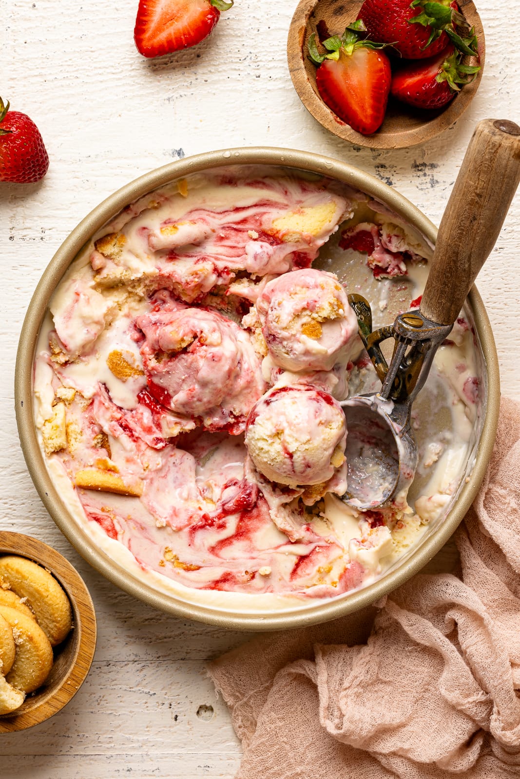 Ice cream in a container with scoops on a white wood table with strawberries and cookies. 