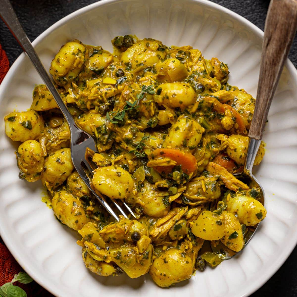 Bowl of curry chicken and gnocchi with a fork + spoon with a cup of juice.