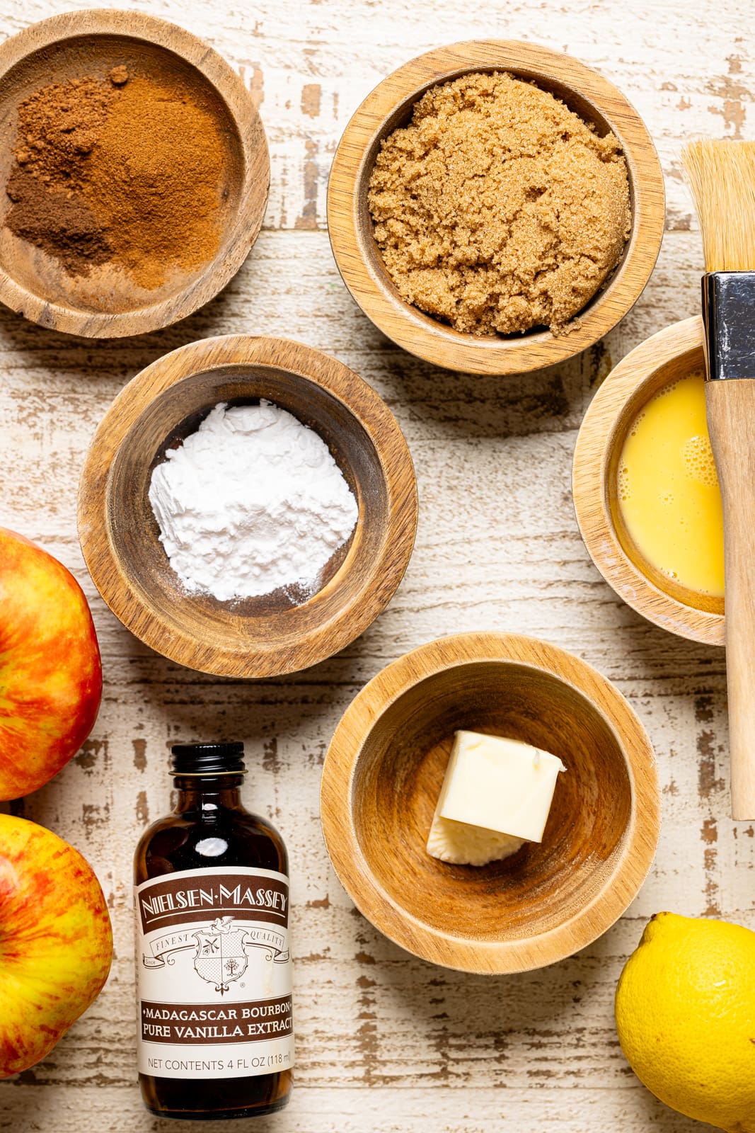 Ingredients on a white wood table including vanilla, brown sugar, spices, apples, butter, and lemon. 