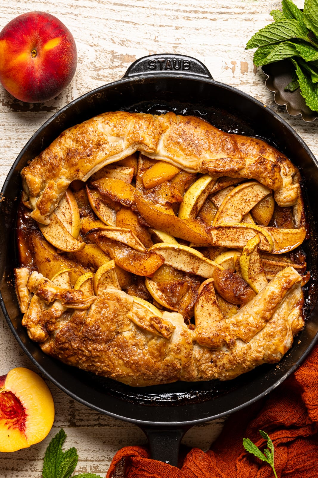 Baked easy peach apple galette with puff pastry in a black skillet with peaches and a red napkin.