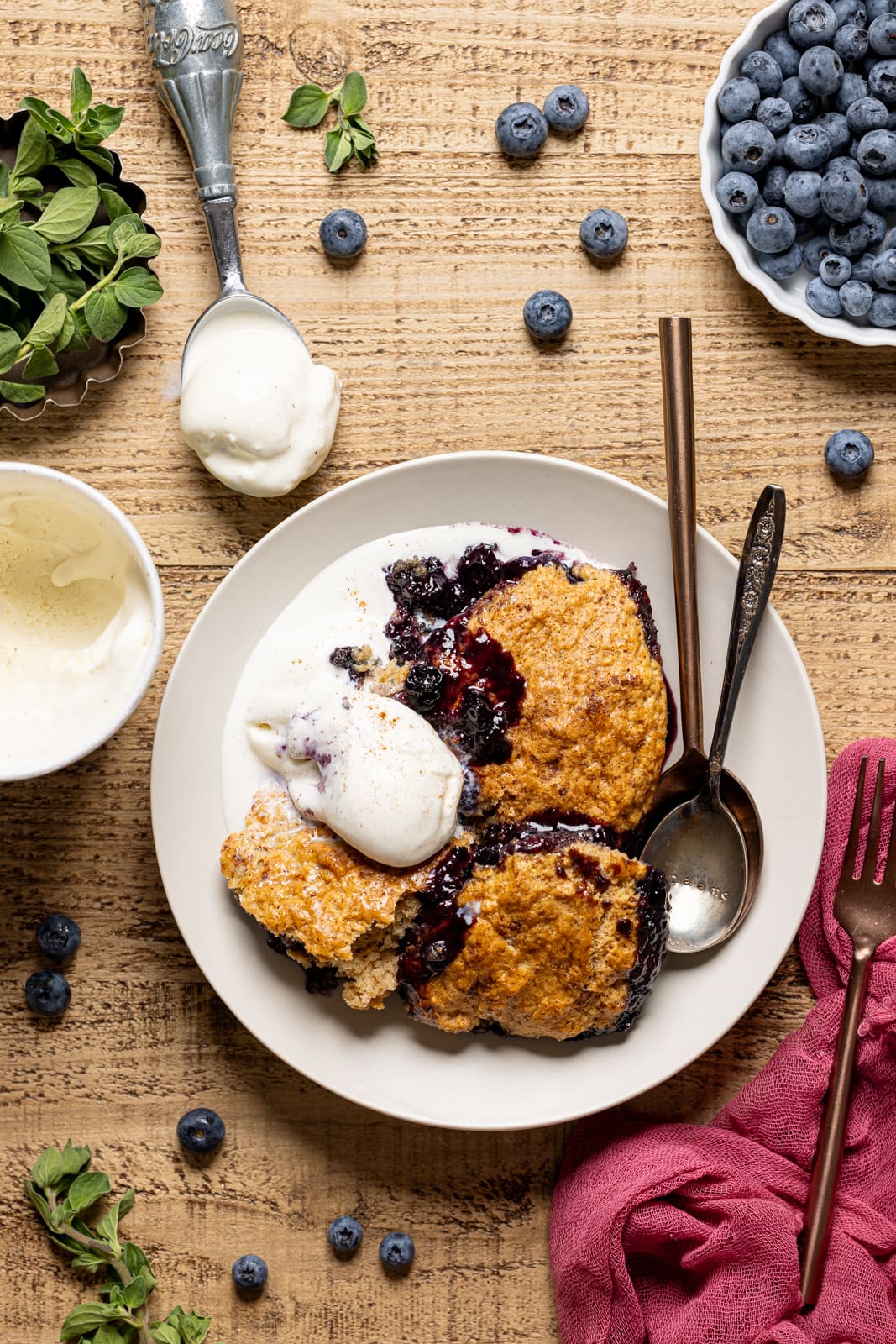 Blueberry cobbler on a plate with two spoons, ice cream scoop, and blueberries.