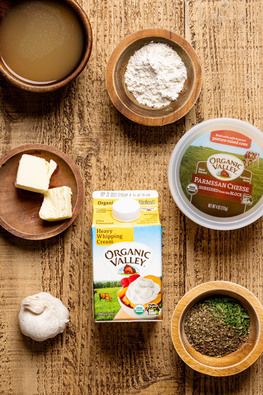 Ingredients on a brown wood table including heavy cream, parmesan cheese, butter, flour, chicken stock, garlic, and herbs + seasonings. 