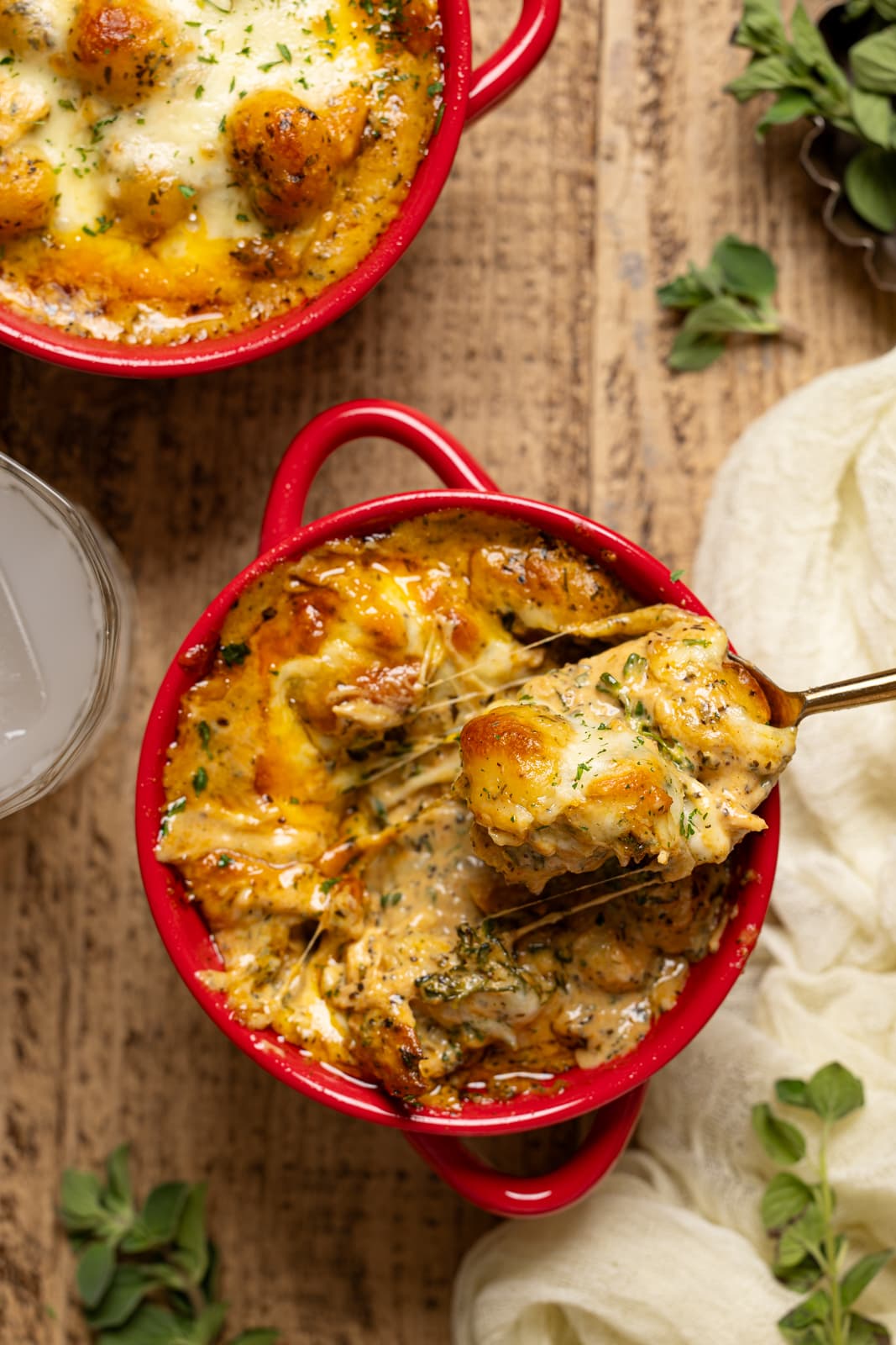 Spoonful of baked gnocchi and cheese in a red bowl, a drink, and herbs.