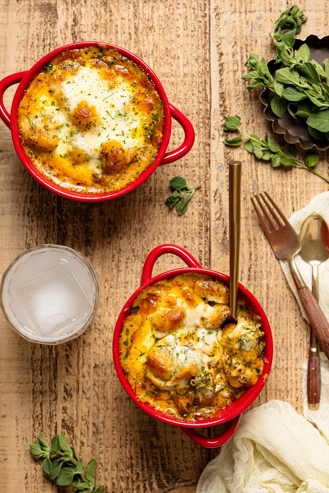 Baked chicken and gnocchi in two red bowls on a brown wood table with a fork, spoon, and drink.