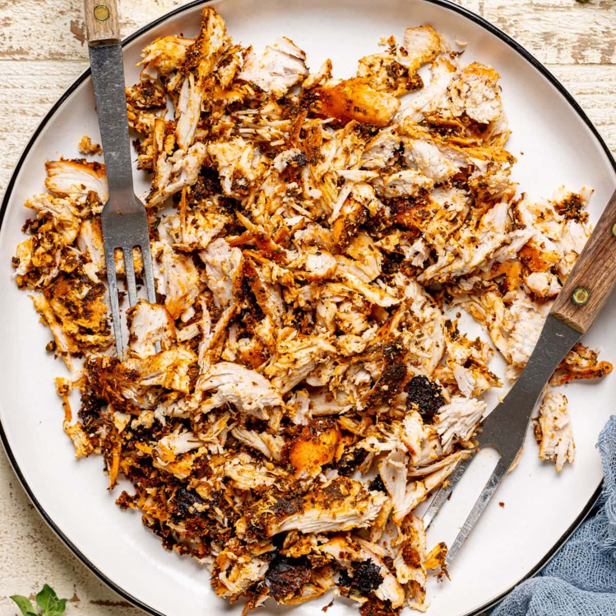 Shredded chicken in a white plate with two forks on a white wood table.