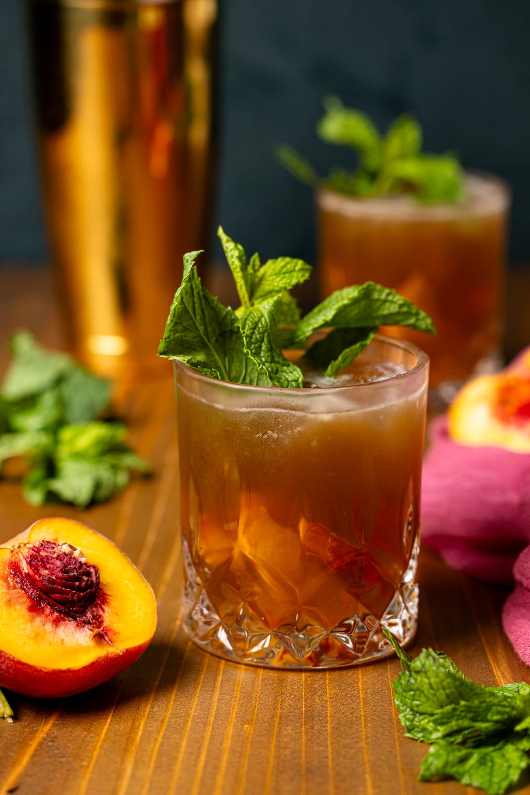 Mokctails in a glass with peaches, mint leaves, and a gold cocktail shaker.