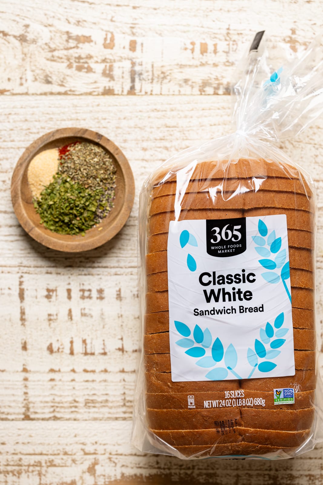 Ingredients on a white wood table including a loaf of sliced white bread and seasoning. 