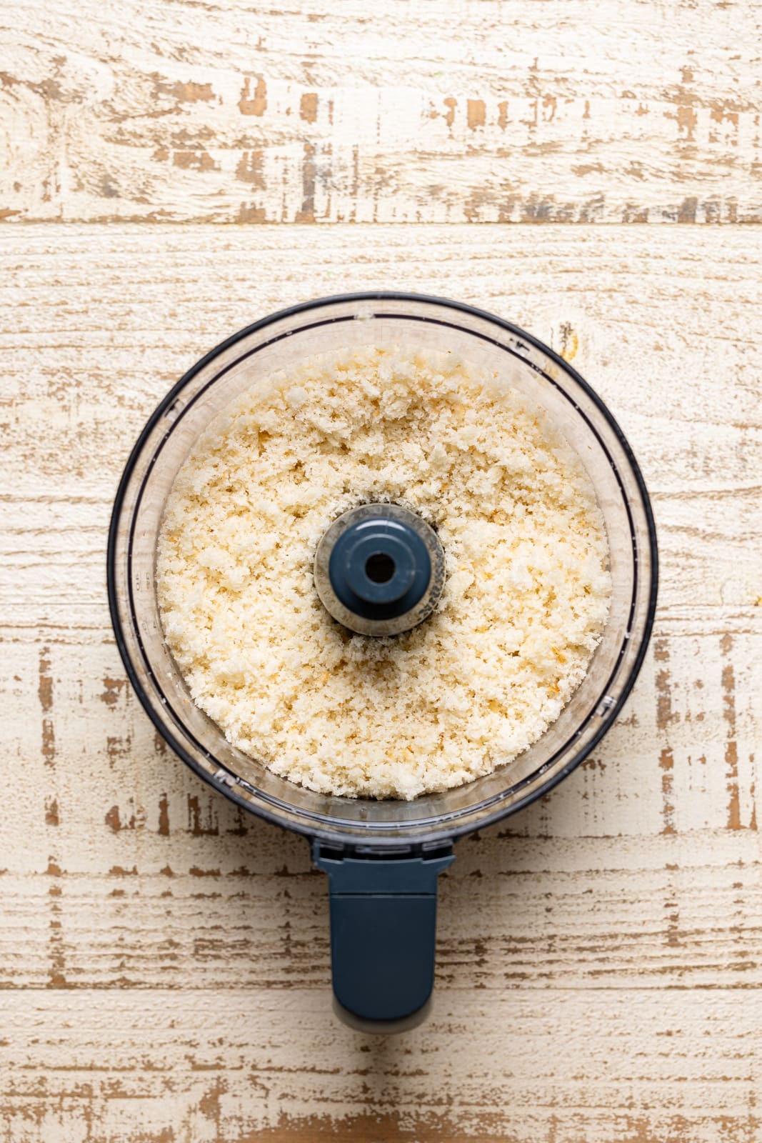 Bread crumbles in a food processor on a white wood table. 