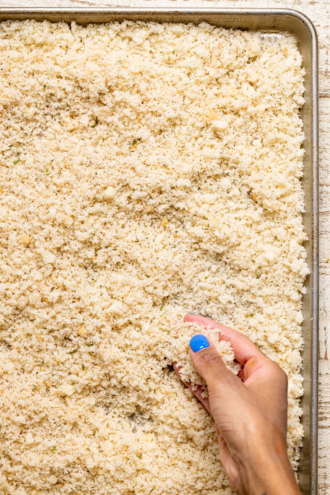 Bread crumbles on a baking sheet with a hand holding crumbles. 