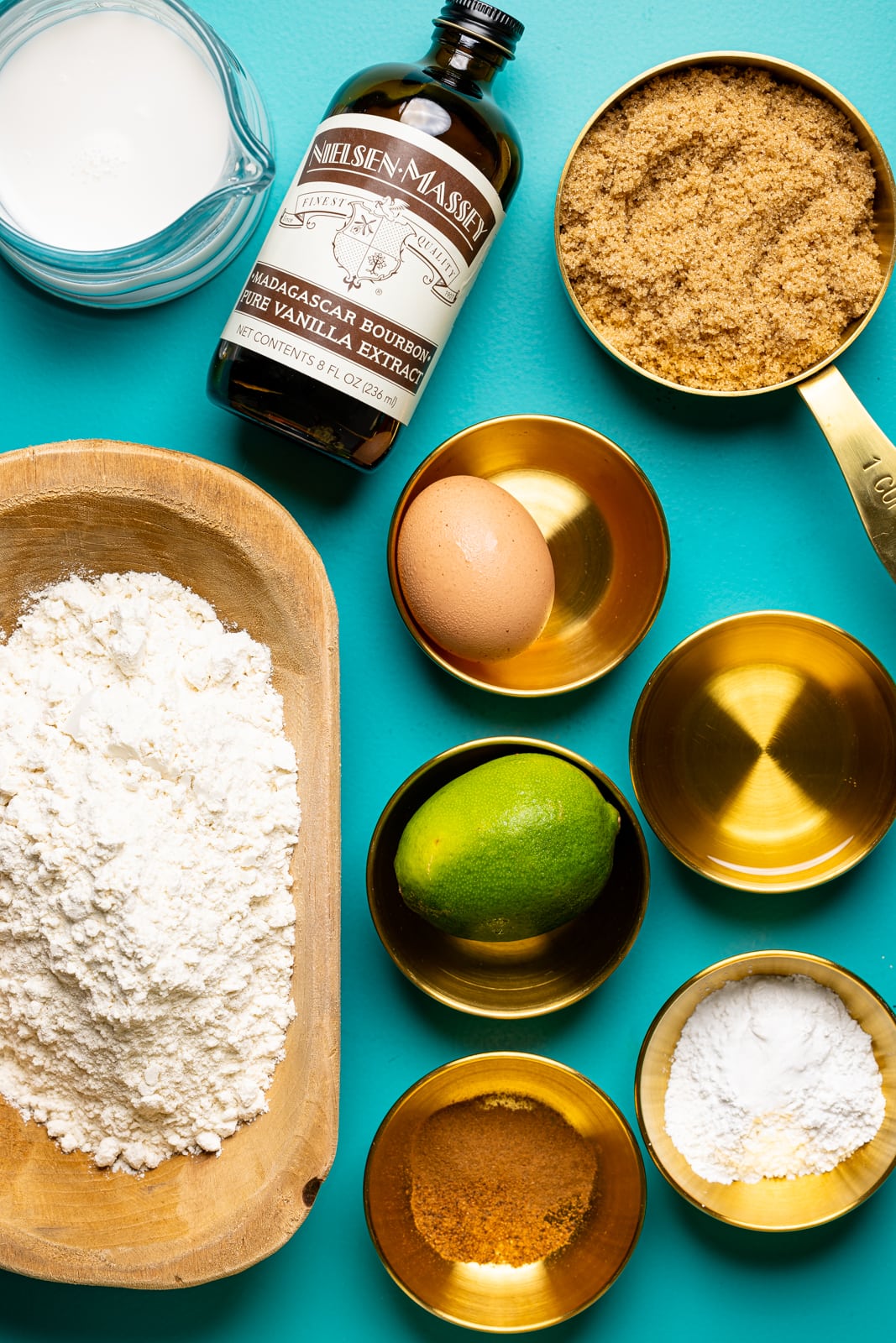 Ingredients on a blue table including egg, vanilla, flour, lime, brown sugar, milk, spices, and oil.