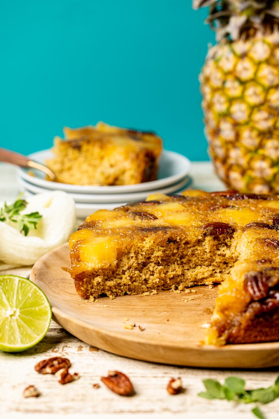 Cake sliced on a brown wood plate with a pineapple in the background and lime and pecans around it.
