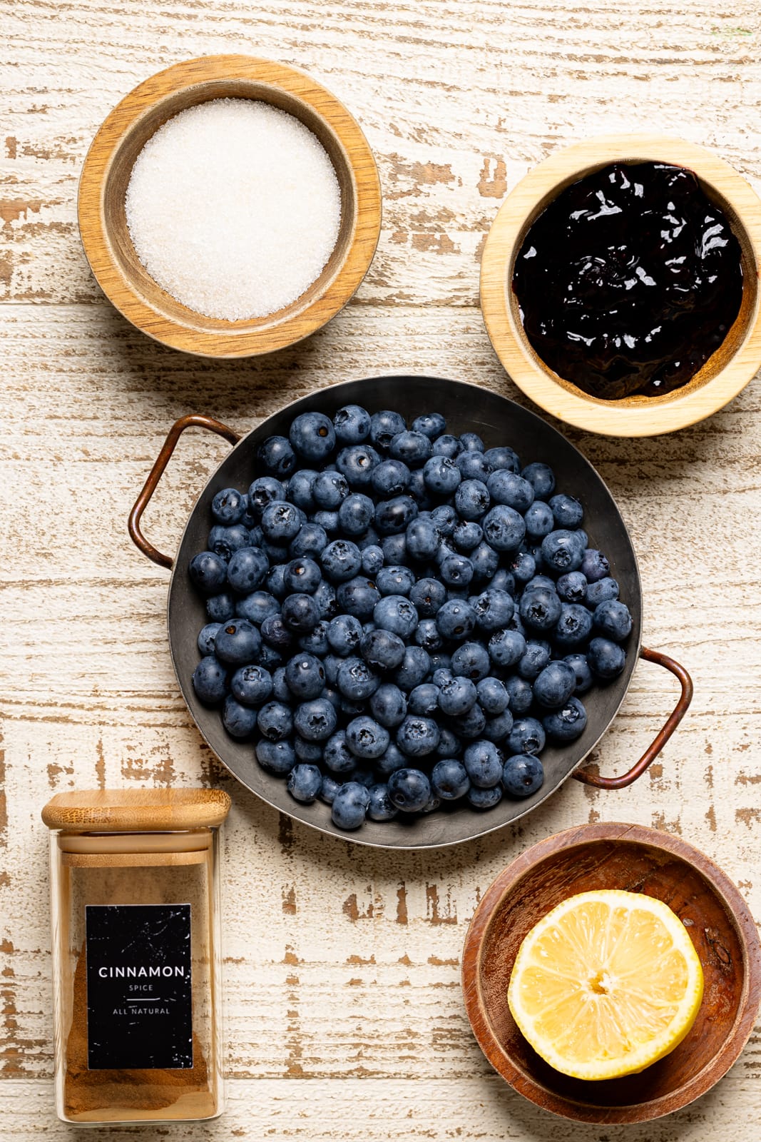 Ingredients on a white wood table including blueberries, sugar, jam, cinnamon, and lemon.