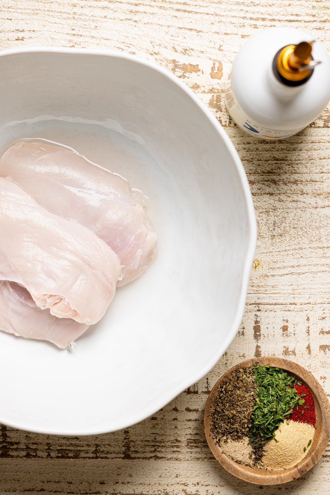 Raw breasts in a white bowl, seasoning, and olive oil on a white wood table.