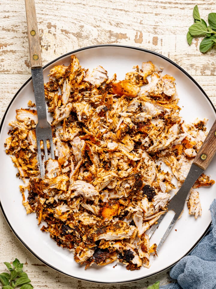 Shredded chicken in a white plate with two forks on a white wood table.