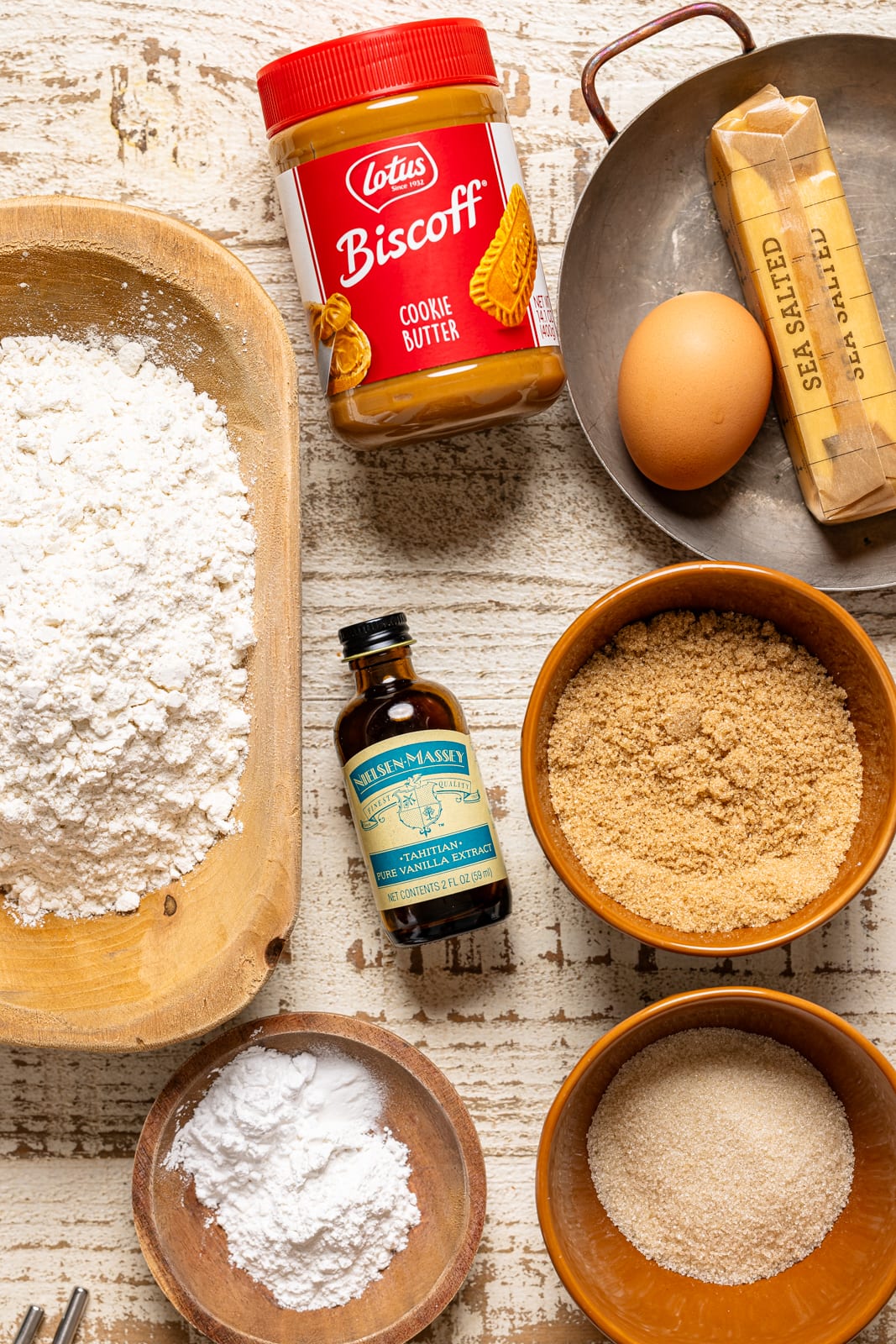 Ingredients on a white wood table including, flour, cookie butter, sugar, vanilla, etc.