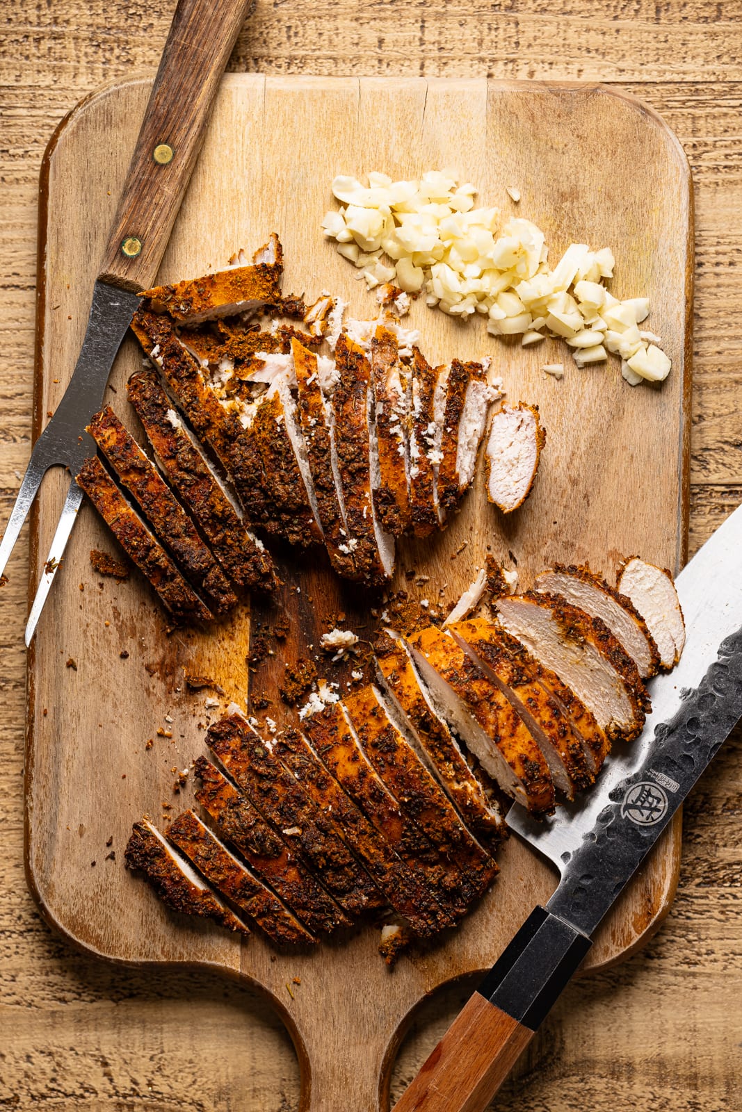 Blackened chicken breasts sliced with garlic on a cutting board with a knife and fork. 