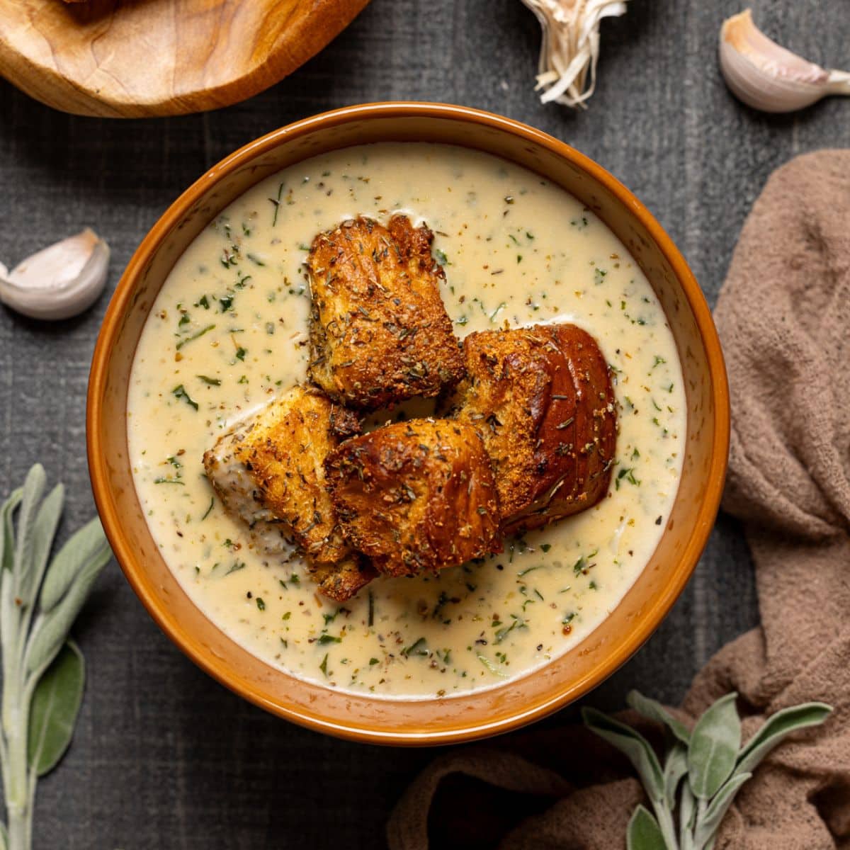 A bowl of soup with garnish of garlic bread, basket of garlic bread, and garlic.