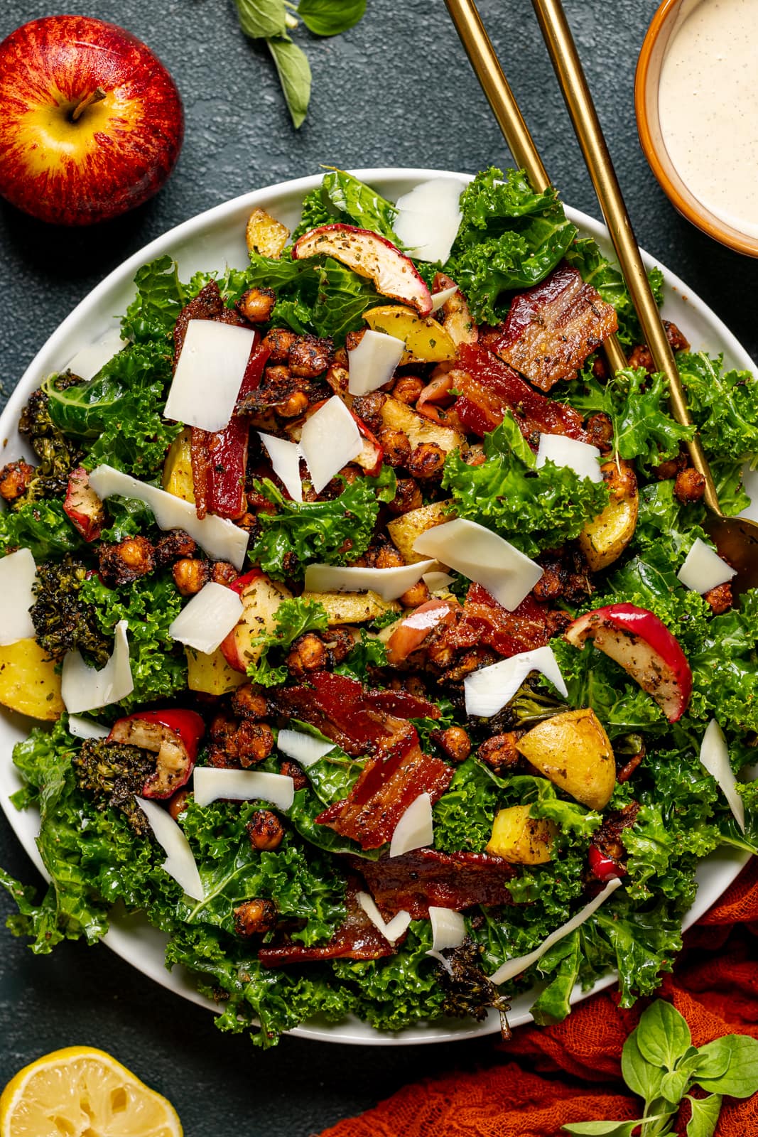 Full kale salad with grated parmesan cheese, two gold serving spoons, and dressing on the side.