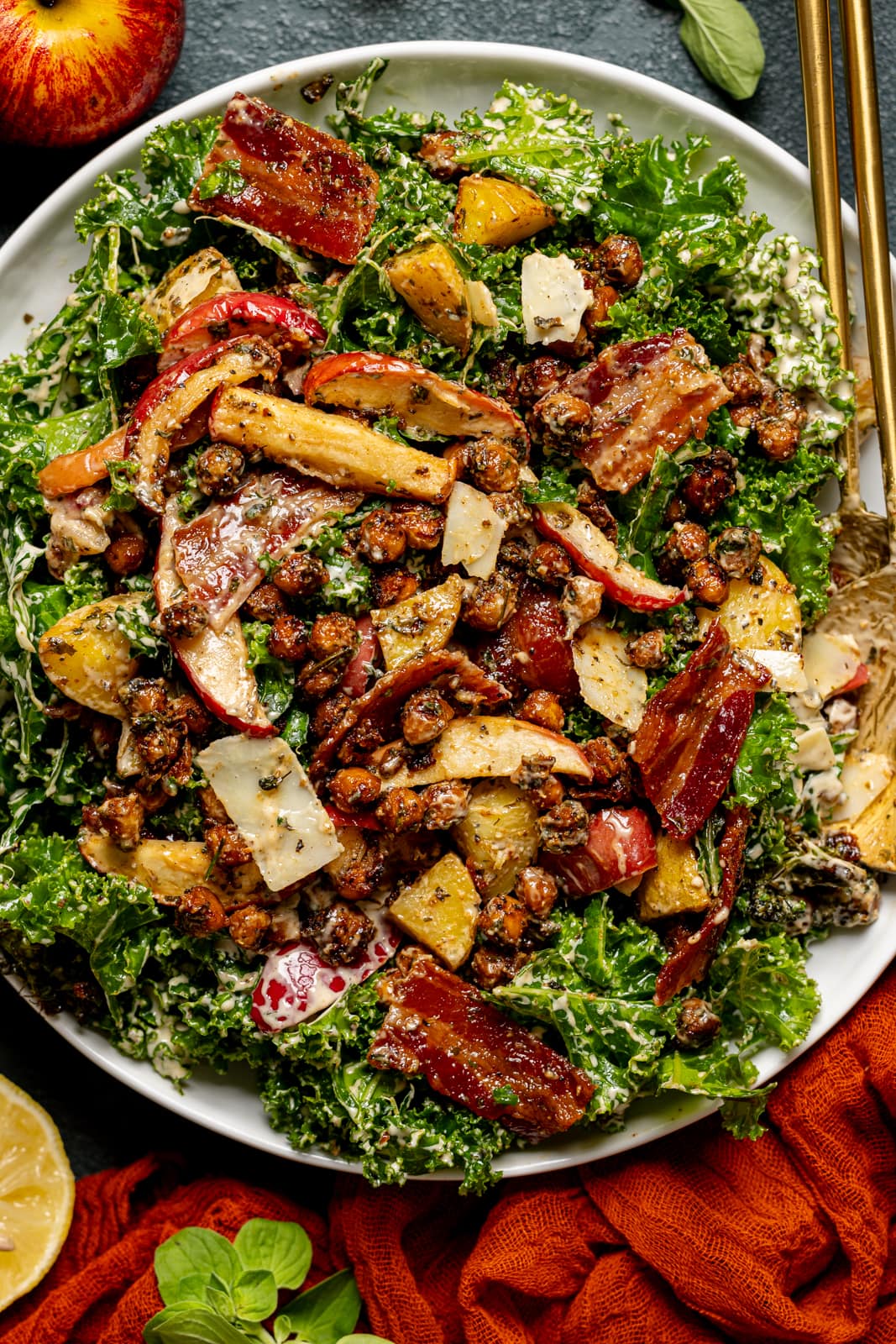 A big bowl of salad with an apple, lemon, and red napkin with gold serving spoons.