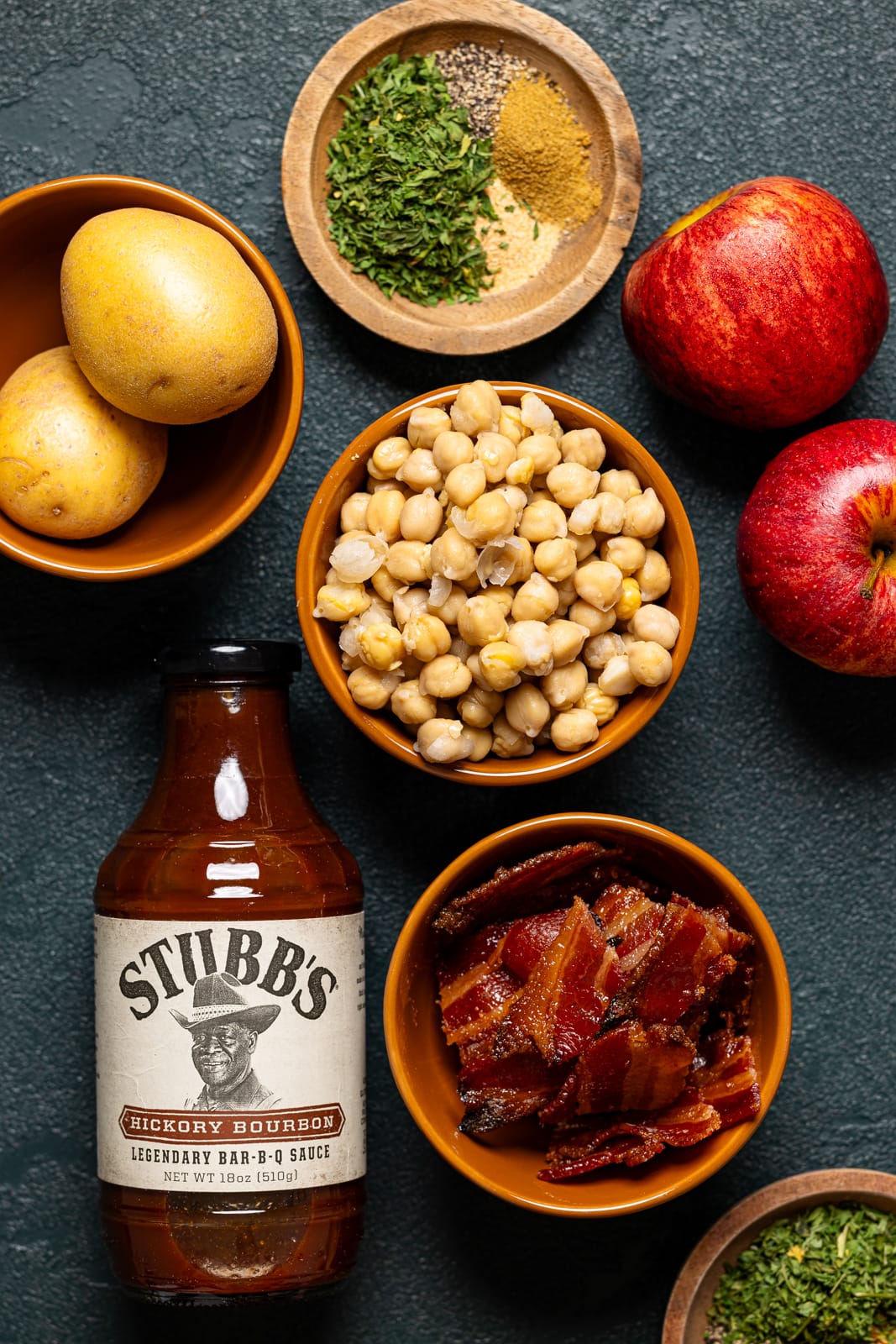 Ingredients on a green table including potatoes, bacon, herbs + seasonings, apples, BBQ sauce, and chickpeas. 