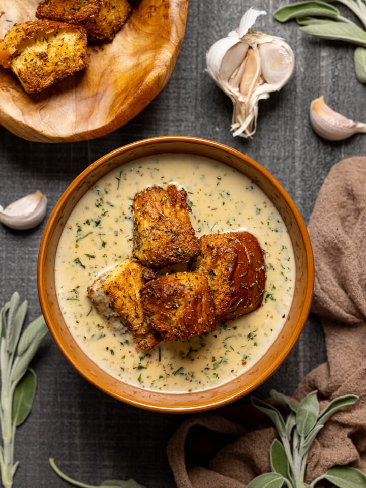 A bowl of soup with garnish of garlic bread, basket of garlic bread, and garlic.