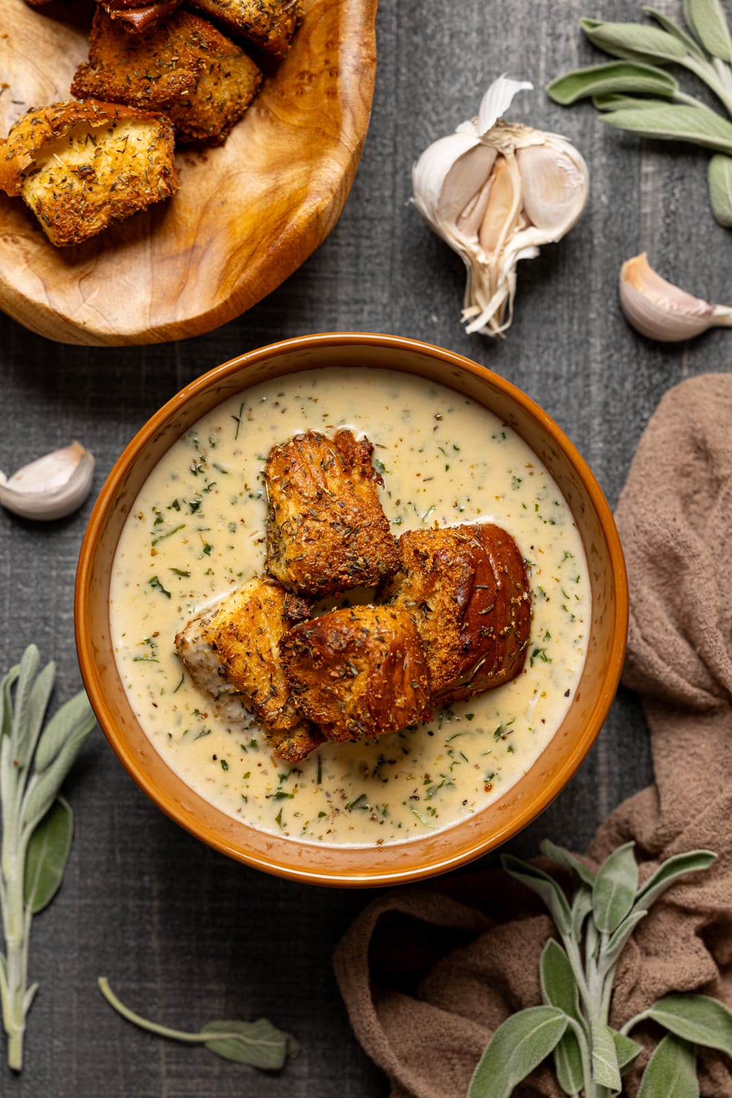 A bowl of soup with garnish of garlic bread, basket of garlic bread, and garlic.