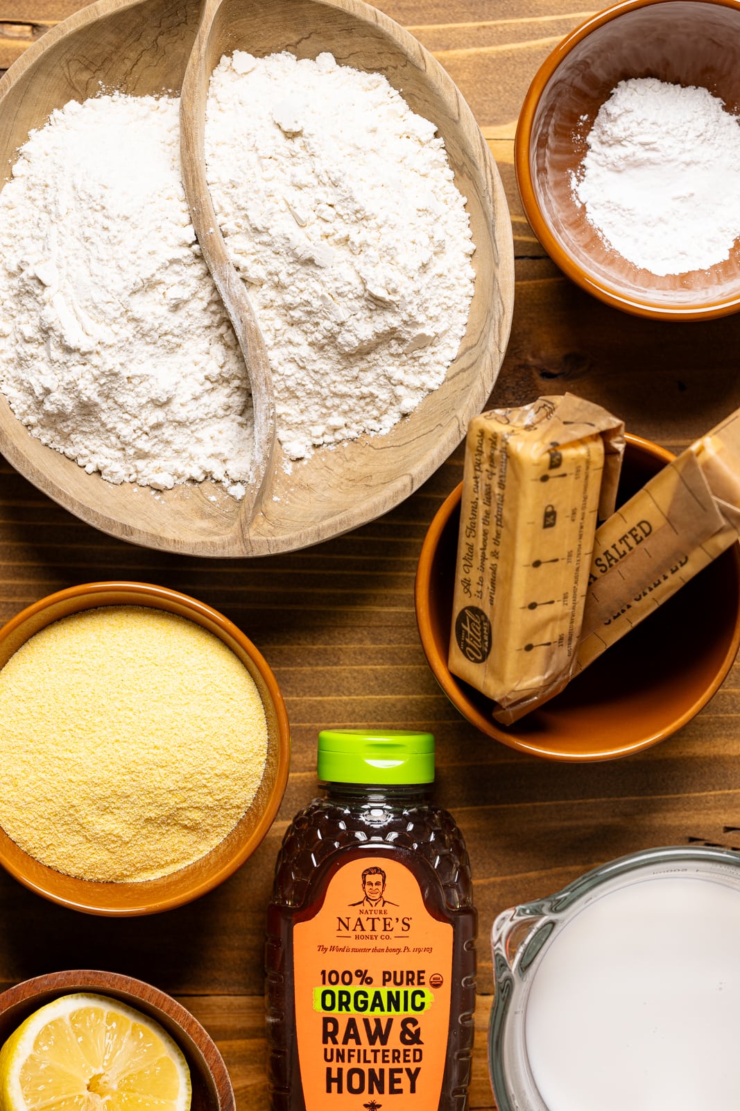 Ingredients on a wood table including flour, cornmeal, honey, butter, and other ingredients. 