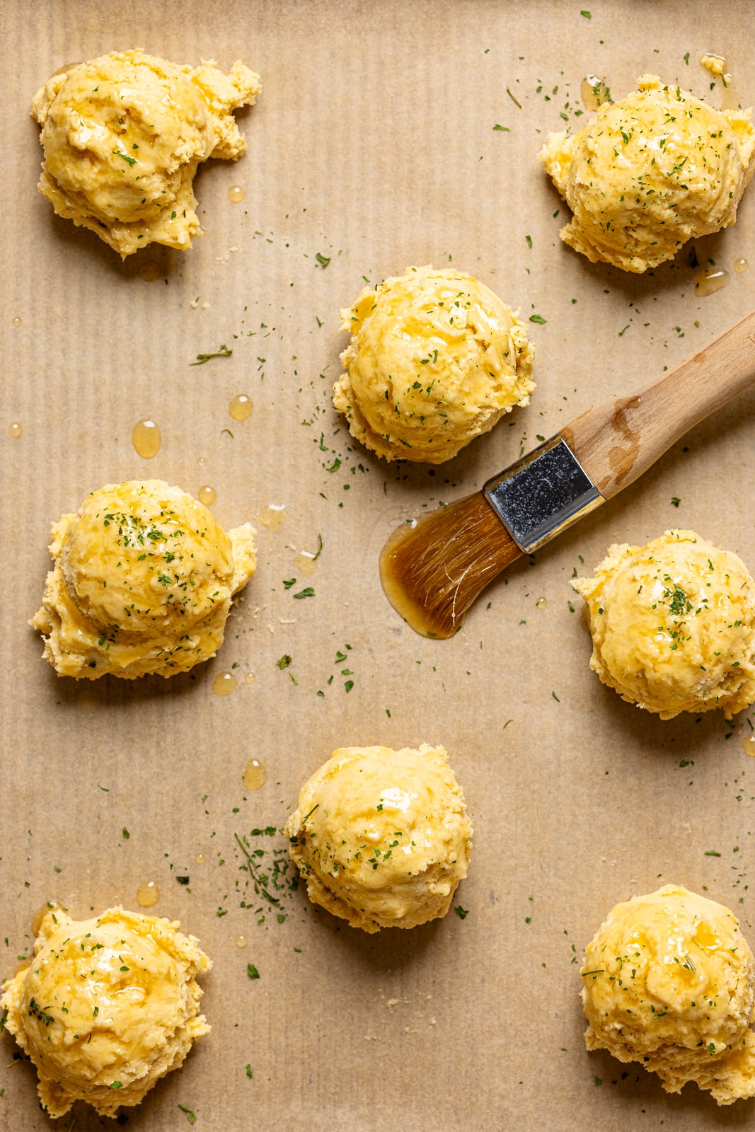 Scooped biscuit dough on a baking sheet with parchment paper with butter brushed on top.