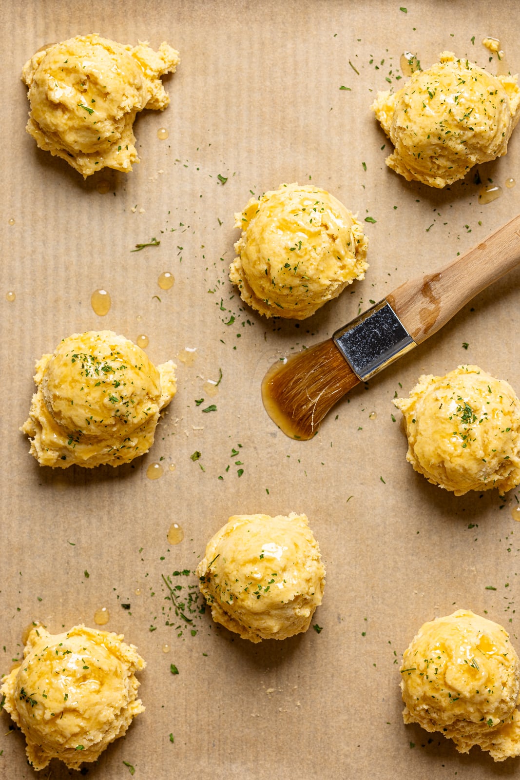 Scooped biscuit dough on a baking sheet with parchment paper with butter brushed on top.