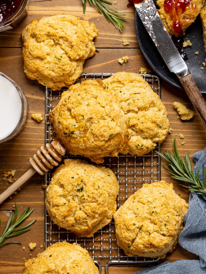 Biscuits on a brown wood table with a glass of milk, honey stick, jam, and plate with a knife.