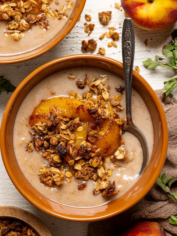 Up close bowl of peach oatmeal with a spoon, fresh peaches, and granola.