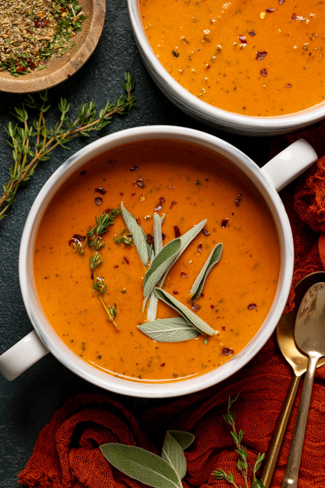 Up close shot of soup in a white bowl with sage and two spoons with a red napkin and grilled cheese. 