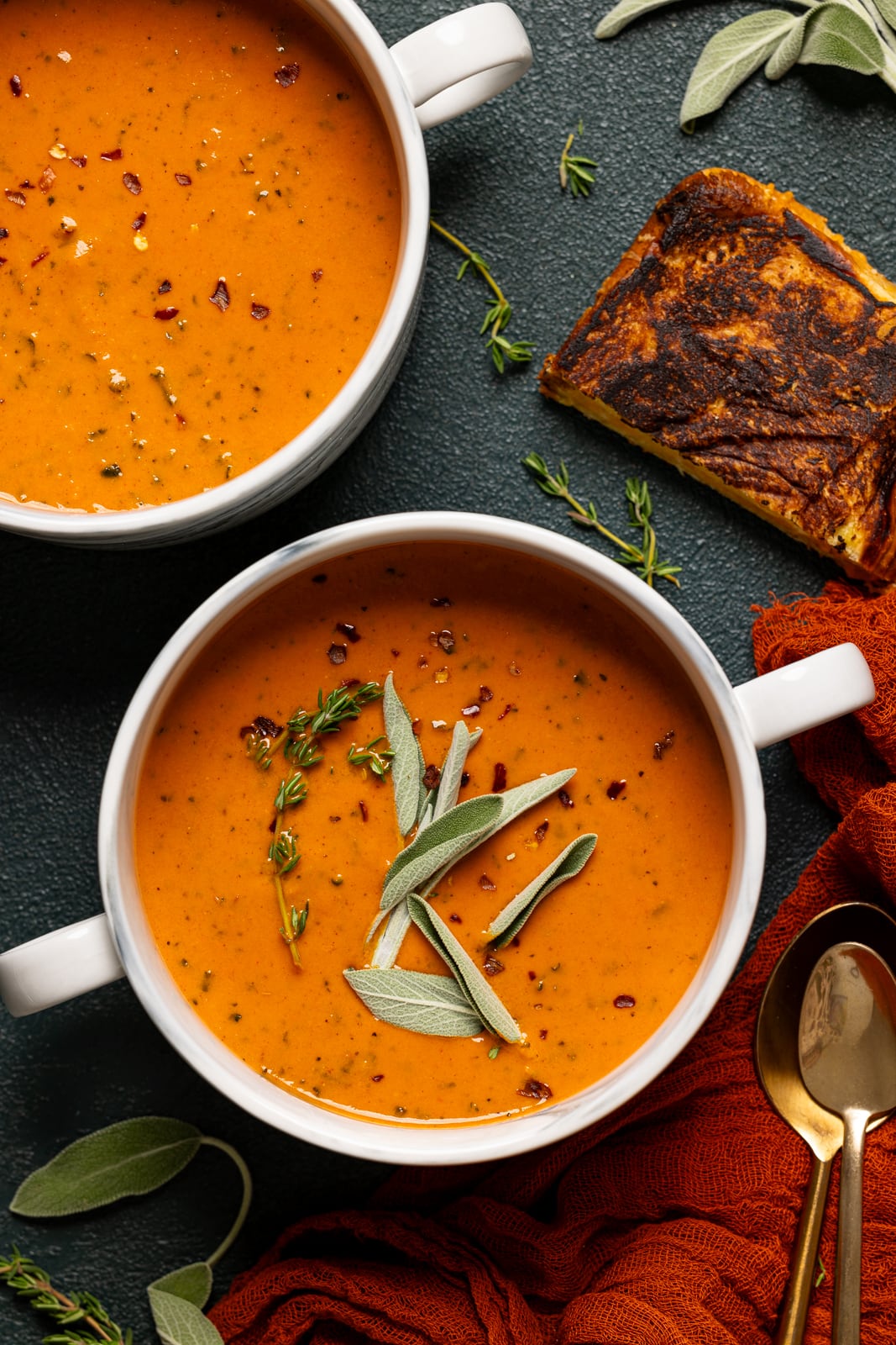 Two bowls of soup with grilled cheese, spoons, and garnish of sage.