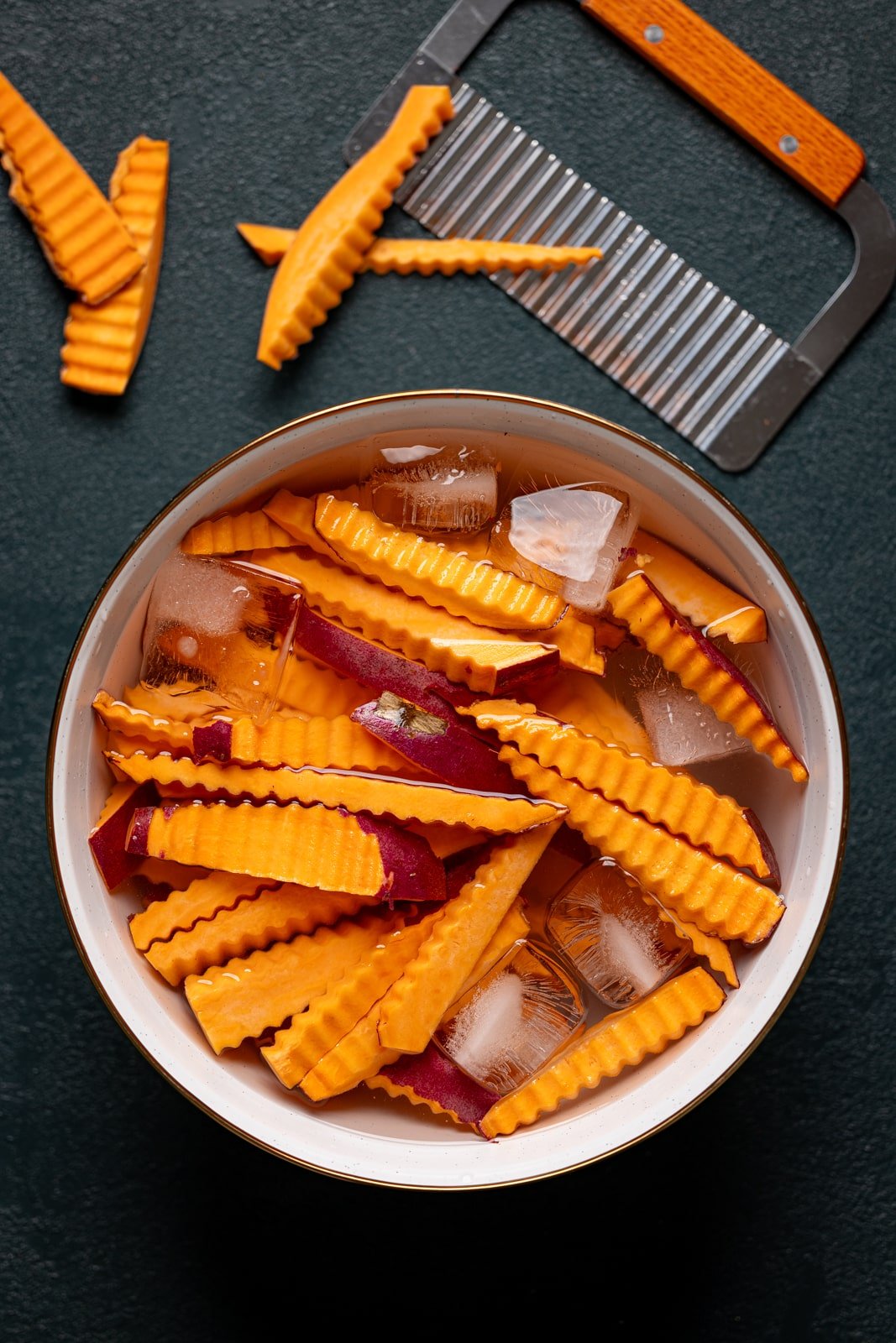 Sweet potatoes cut into fries with a cutter and in a white bowl with water + ice. 