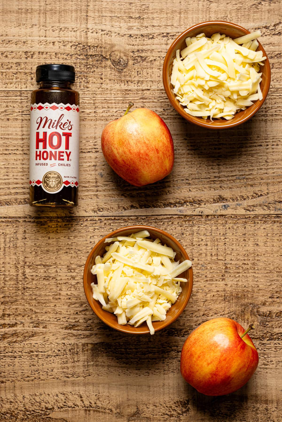 Apples, hot honey, and cheddar cheese in bowls on a brown wood table.
